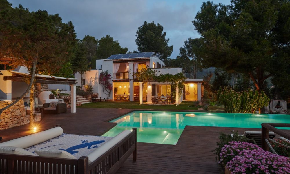 Evening view of a luxurious villa with a pool, outdoor seating, and lush greenery, illuminated by warm lights.