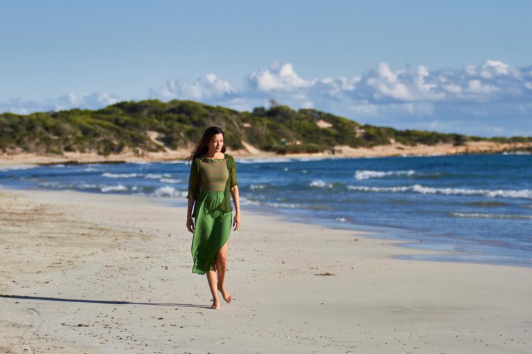 Woman in a green dress walking on a beach with the sea and hills in the background.