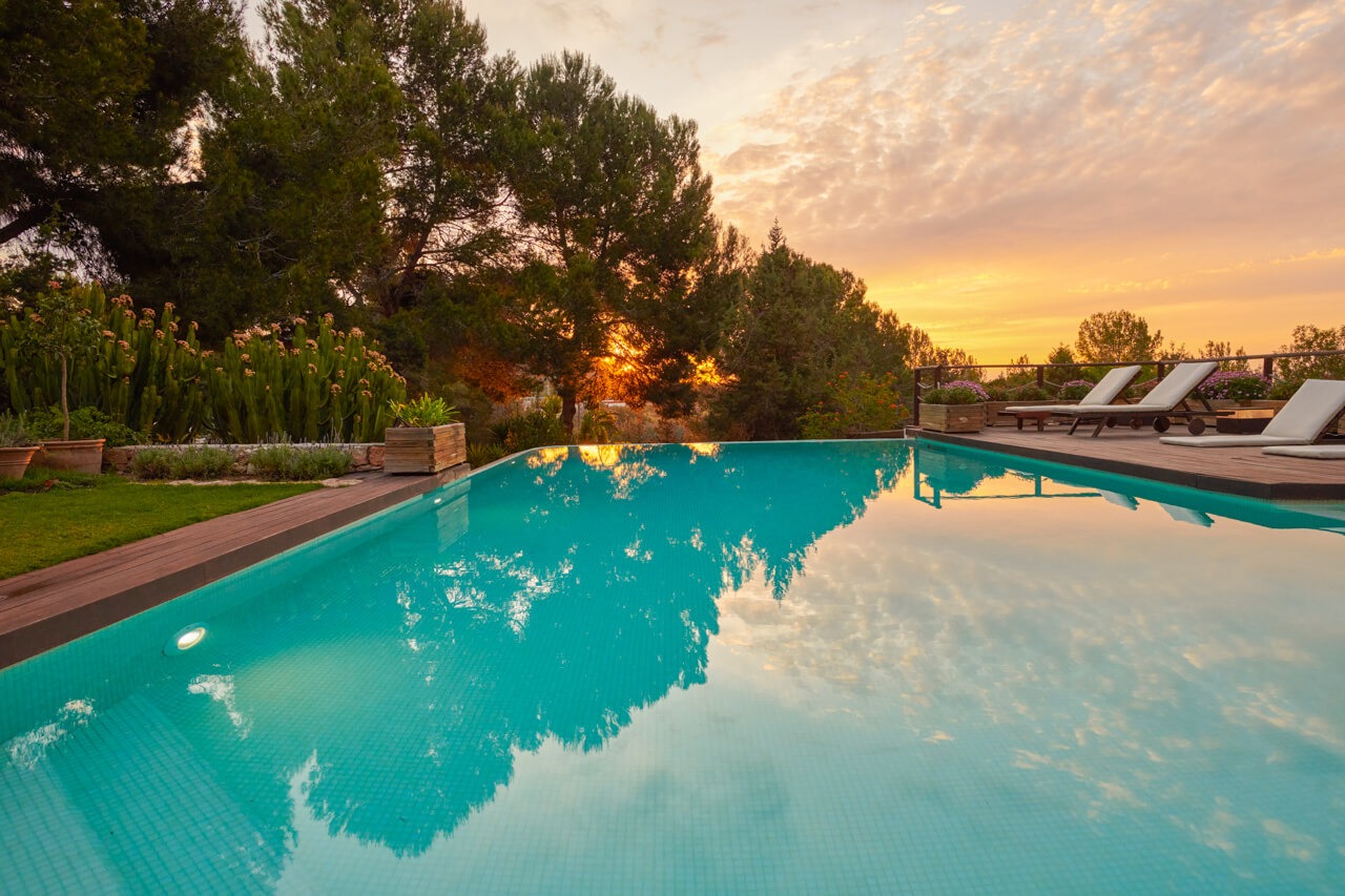 Evening view of a luxurious villa with a pool, outdoor seating, and lush greenery, illuminated by warm lights.